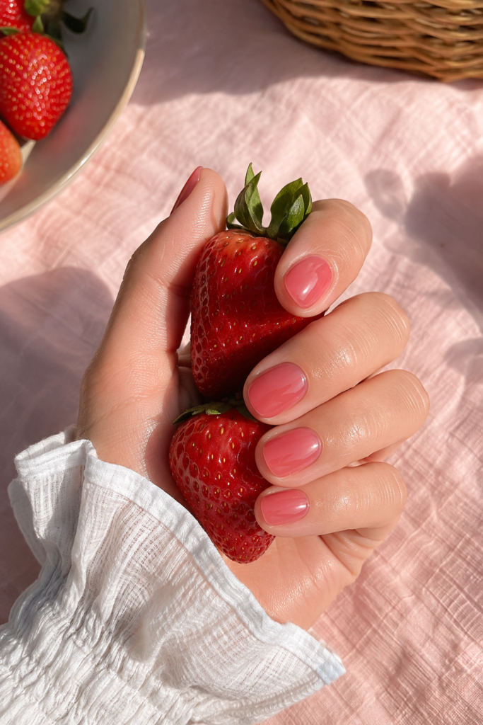 Short strawberry-pink jelly nails with glossy, glass-like shine, hand holding fresh strawberries over a pale pink picnic cloth.