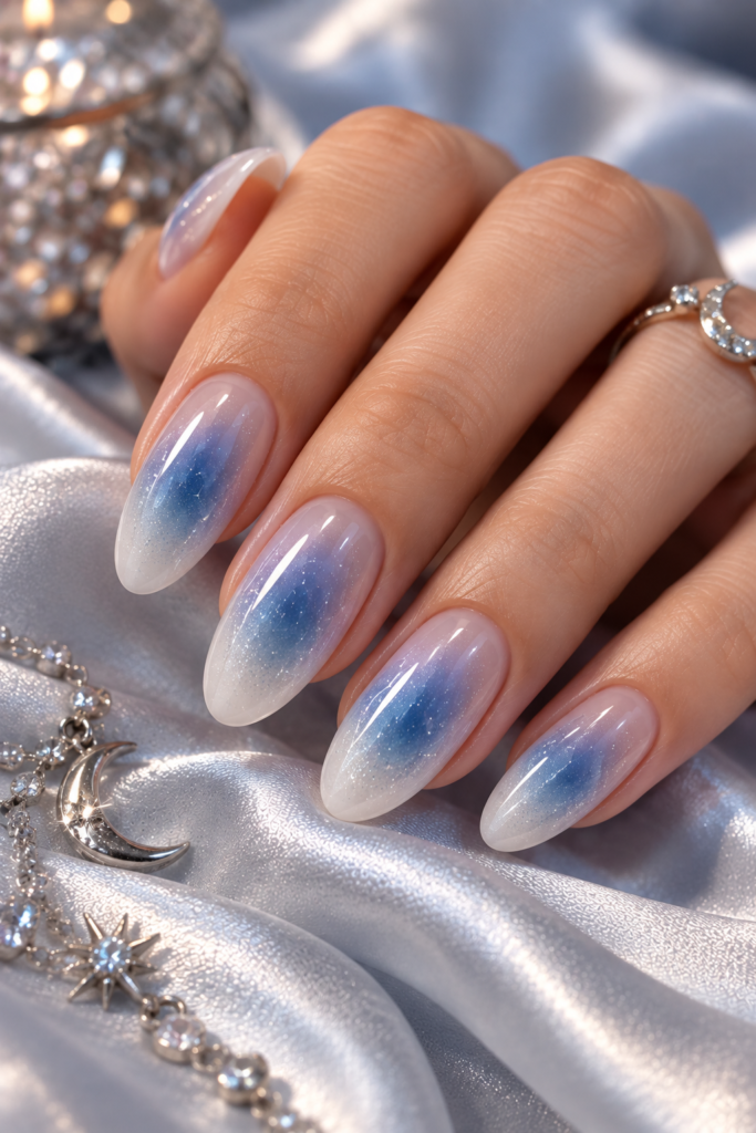 A hand with long, almond-shaped nails featuring a blue and white aura gradient design, resting on silver satin fabric next to a crystal-encrusted box and a moon and star necklace.