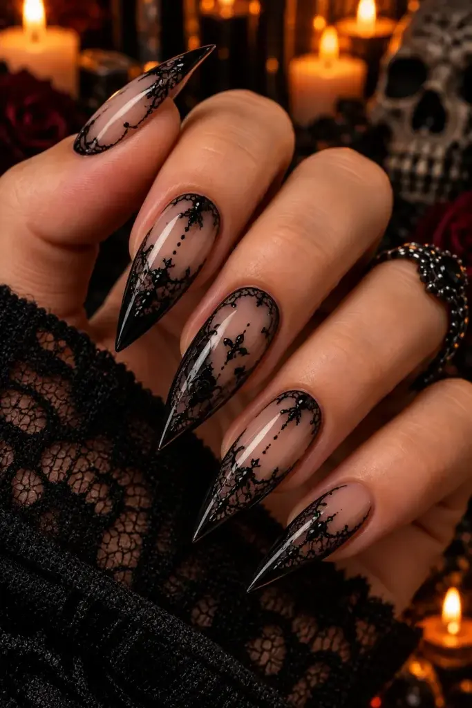 A hand with long, pointed stiletto nails featuring a nude base and intricate black lace patterns, set against a dark background with candles and a skull.