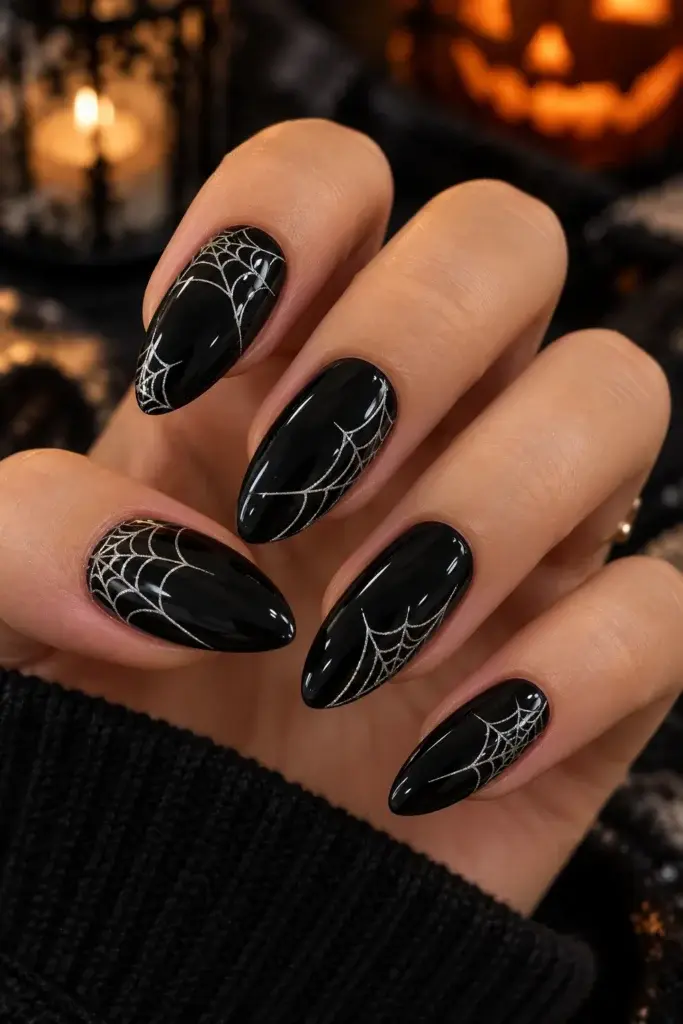 A hand with long, almond-shaped black nails featuring silver spider web designs, set against a dark background with a lit candle.