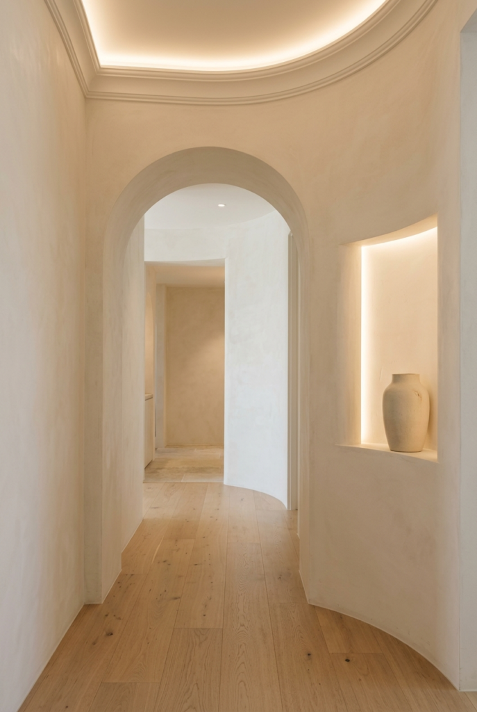 A minimalist hallway featuring curved plaster walls, light oak flooring, and a recessed niche with a ceramic vase illuminated by hidden LED lighting.