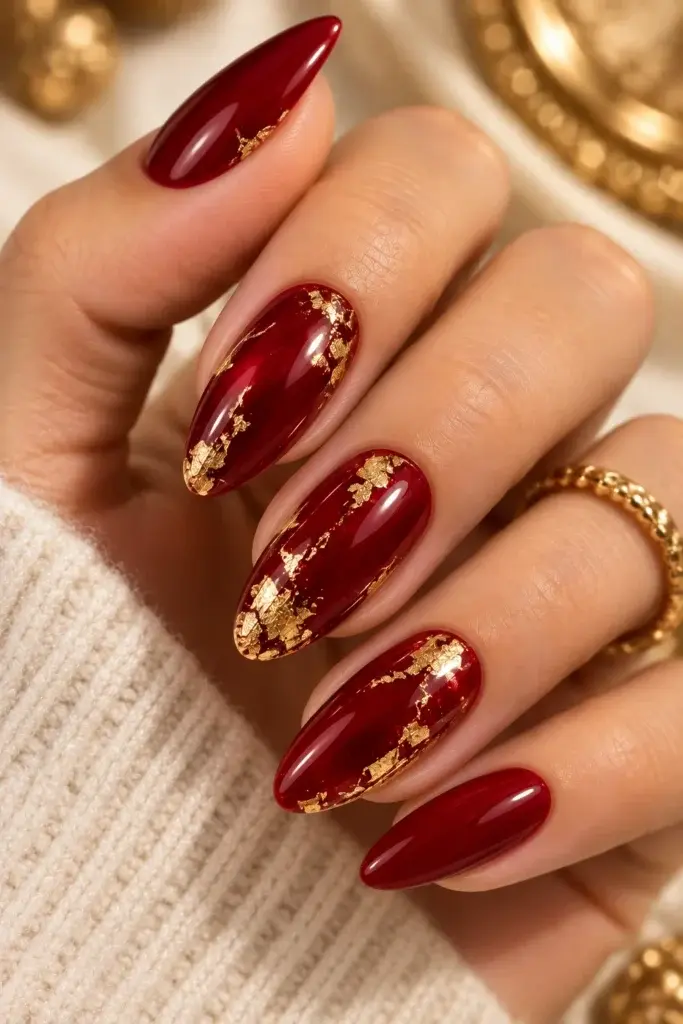 A close-up of long, almond-shaped nails painted in a deep red shade, featuring gold foil accents on the middle fingers, set against a cream knit sweater.
