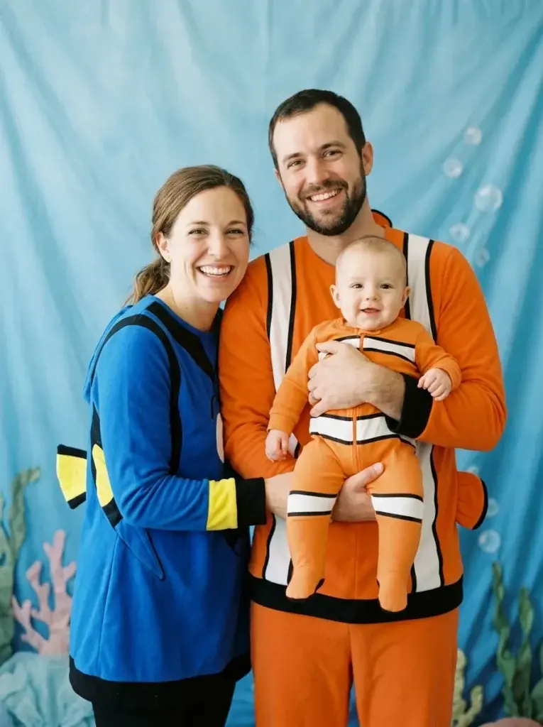 A family of three wearing Finding Nemo Halloween costumes, with the father holding a baby in a matching orange clownfish onesie.