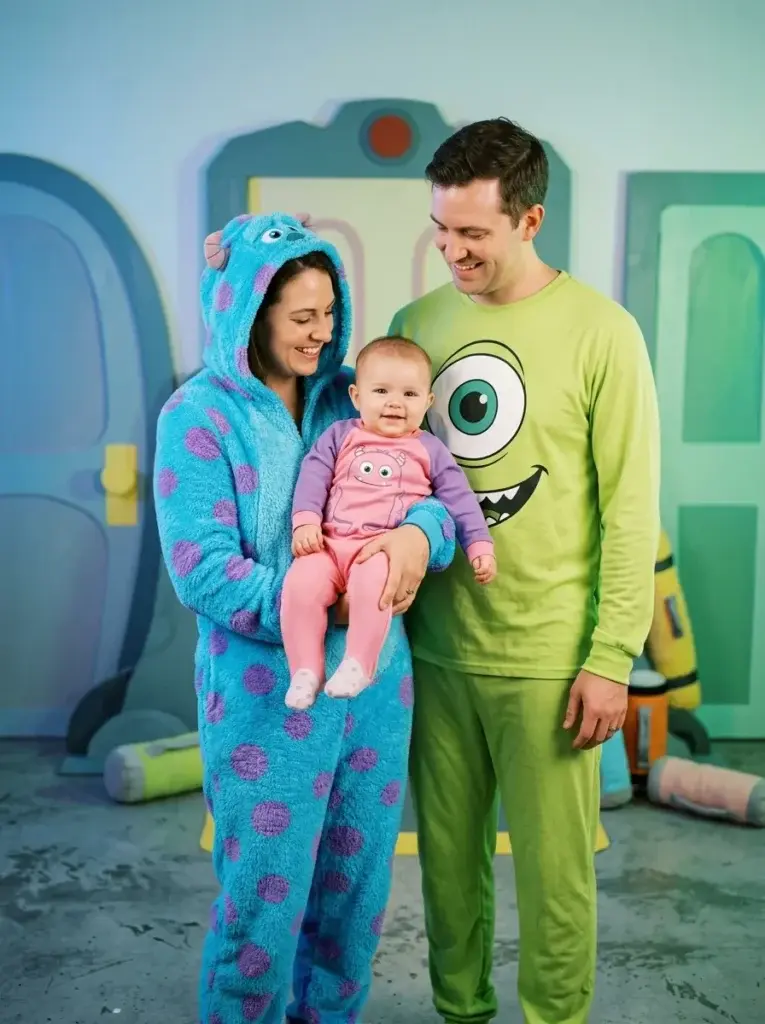 A family of three wearing matching Monsters, Inc. pajama costumes, with the mother in a blue Sulley onesie holding a baby in a pink onesie, and the father in a green Mike Wazowski shirt.