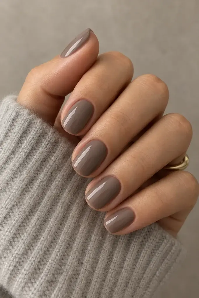 A hand with short, almond-shaped nails painted in a glossy, muted taupe shade, resting against a ribbed grey knit sleeve with a thin gold ring on the ring finger.