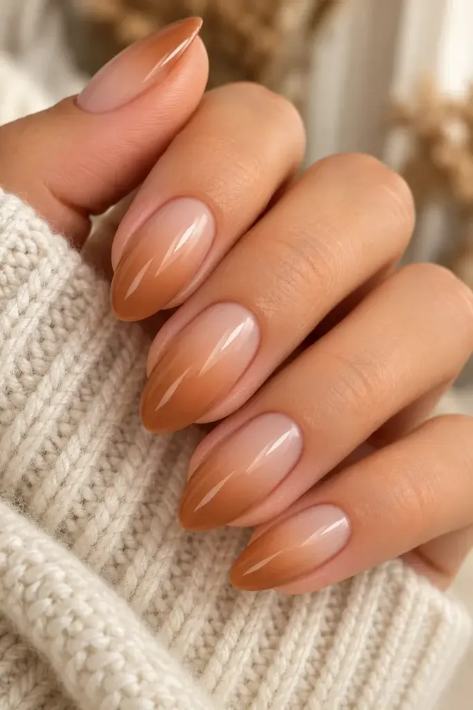 Close-up of almond-shaped nails with a smooth nude to caramel ombré finish, resting against a cream-colored knit sweater sleeve.