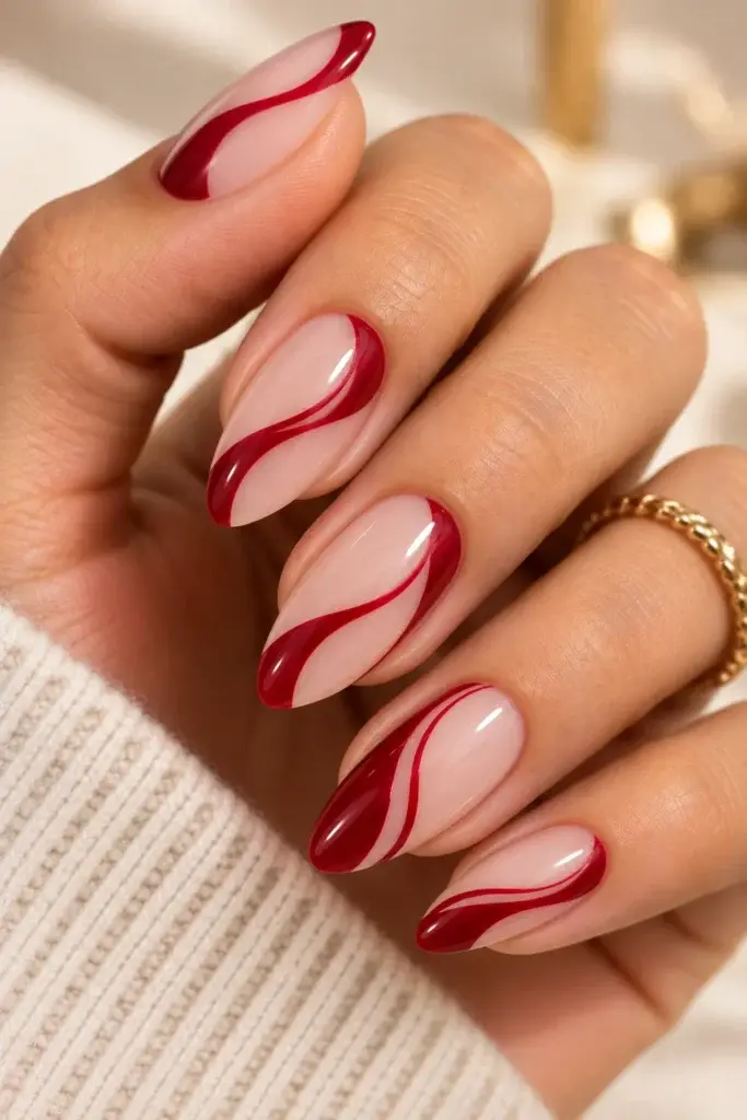 A close-up of long, almond-shaped nails featuring a glossy light pink base with bold, wavy dark red accents, worn on a hand with a gold ring.