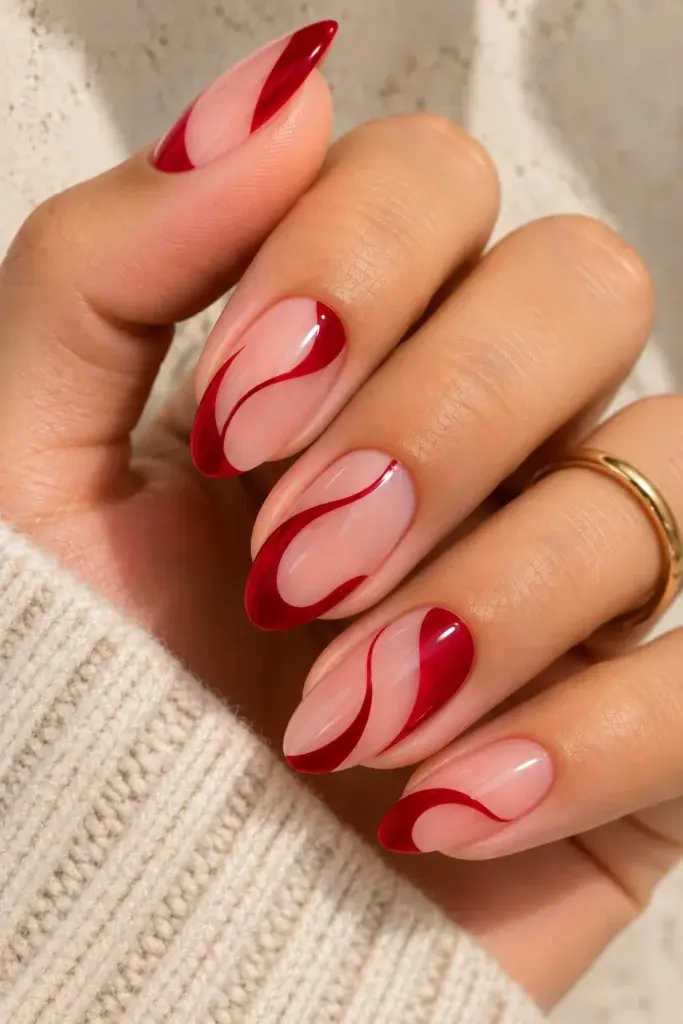 A close-up of almond-shaped nails featuring a glossy pink base with bold, wavy red accents, worn on a hand with a gold ring and cream sweater.