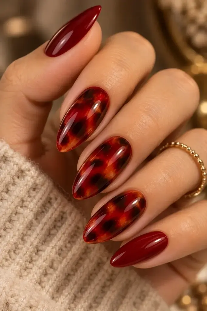 A hand with long almond-shaped nails featuring a mix of solid deep red and red tortoiseshell patterns, wearing a gold beaded ring.