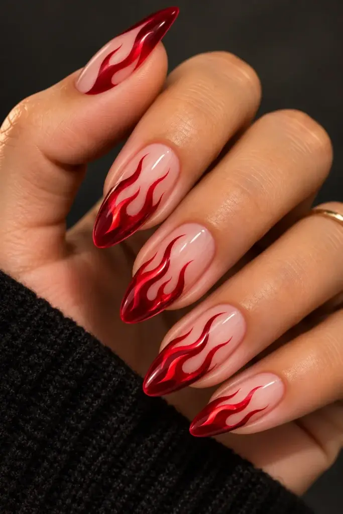 Close-up of long, almond-shaped nails featuring a red flame design on a pale pink base, shown on a hand wearing a black knit sleeve.