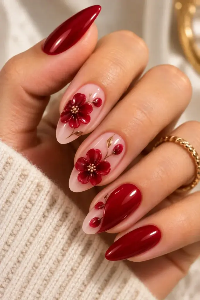 A close-up of long almond-shaped nails featuring a mix of solid deep red polish and sheer pink bases decorated with 3D red flowers and gold accents.