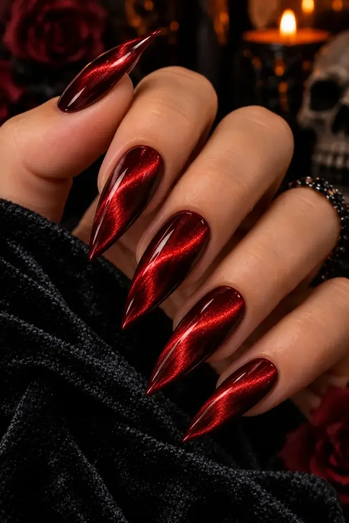 A hand with long, pointed red nails featuring a metallic finish, set against a dark background with a candle and skull.